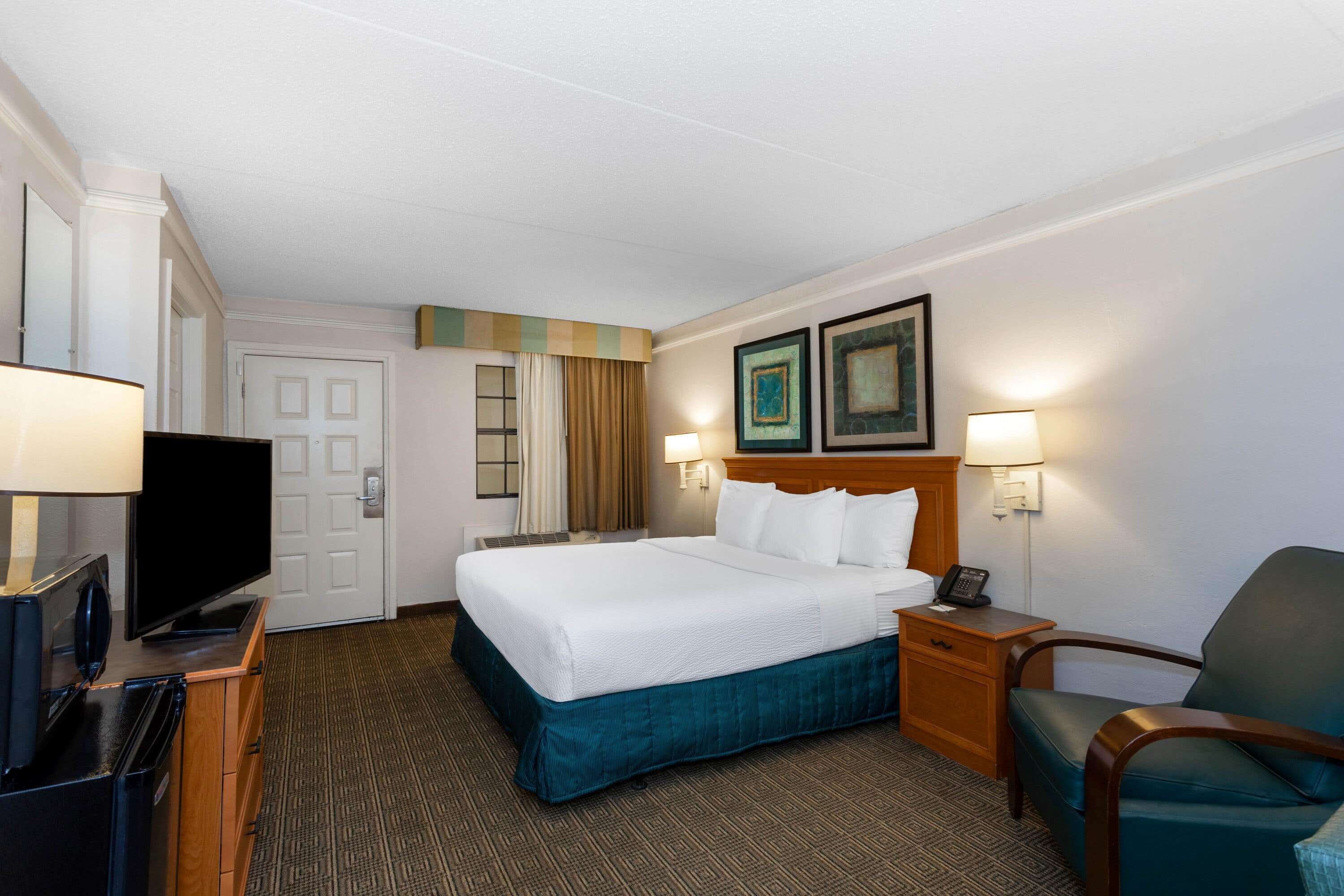Quarto de hotel com uma cama grande, televisão, escrivaninha com abajur, cadeira e duas imagens emolduradas na parede.