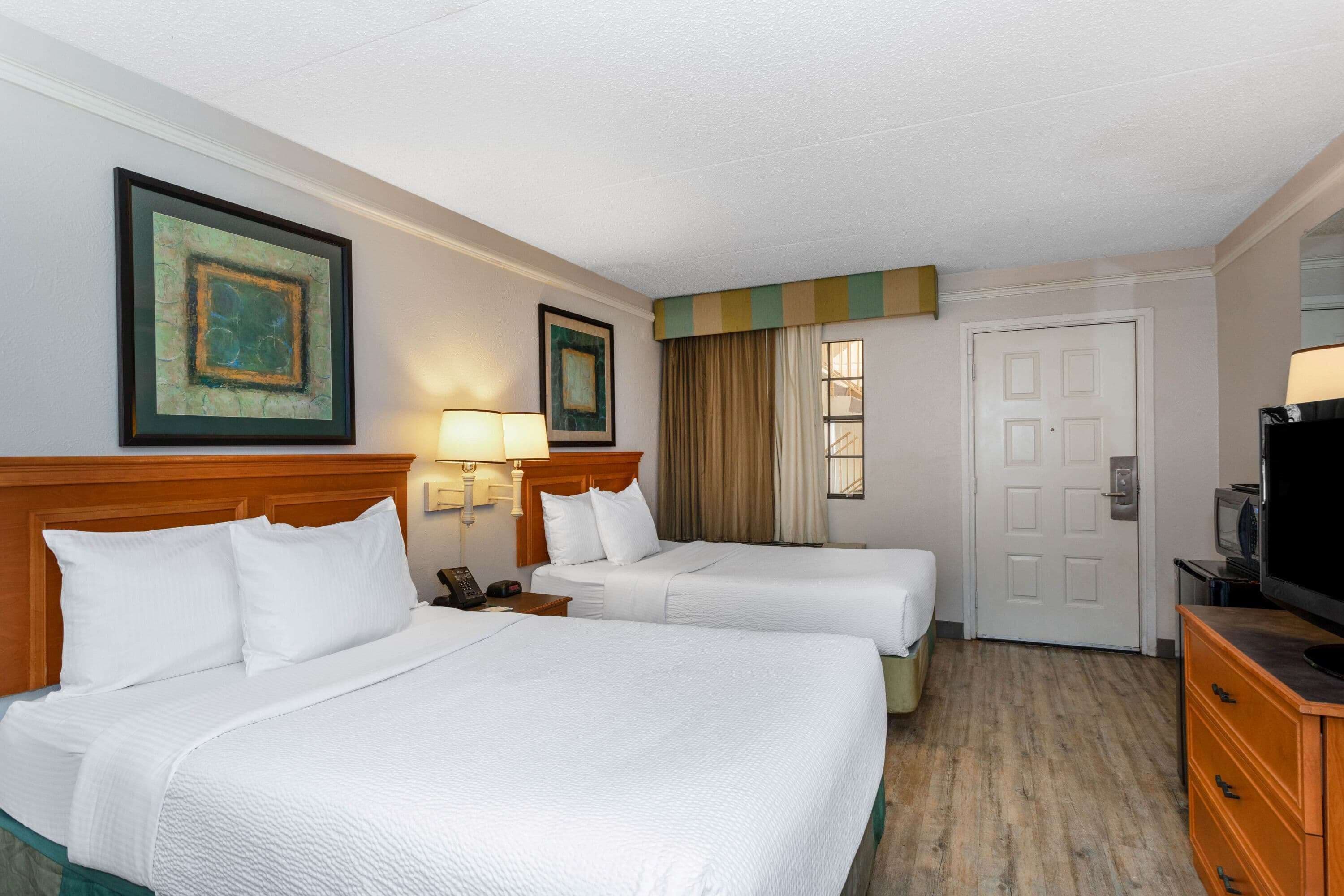 A hotel room with two beds, a wooden headboard, a bedside table with a lamp, a television, and a door.
