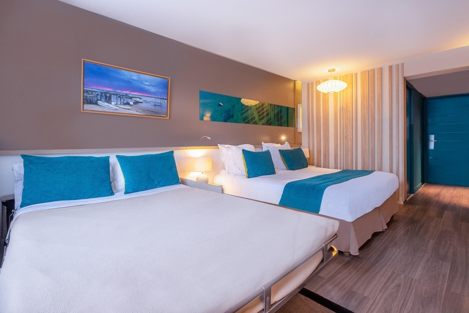 A hotel room with two beds, a painting, a lamp, and a view of the outside.