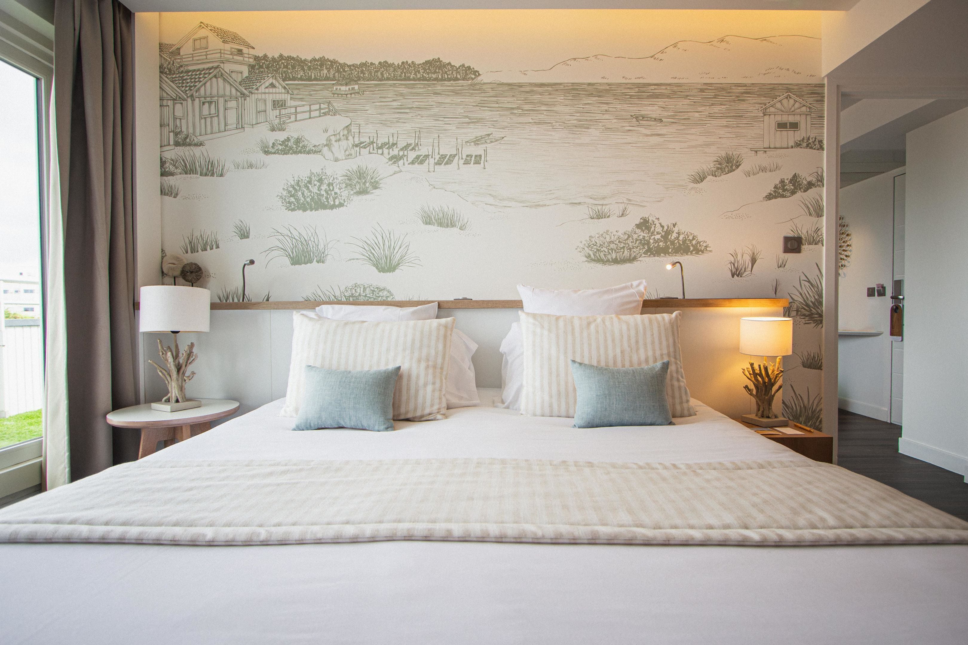 A hotel room with a large bed, two bedside lamps, and a mural of a coastal scene on the wall.
