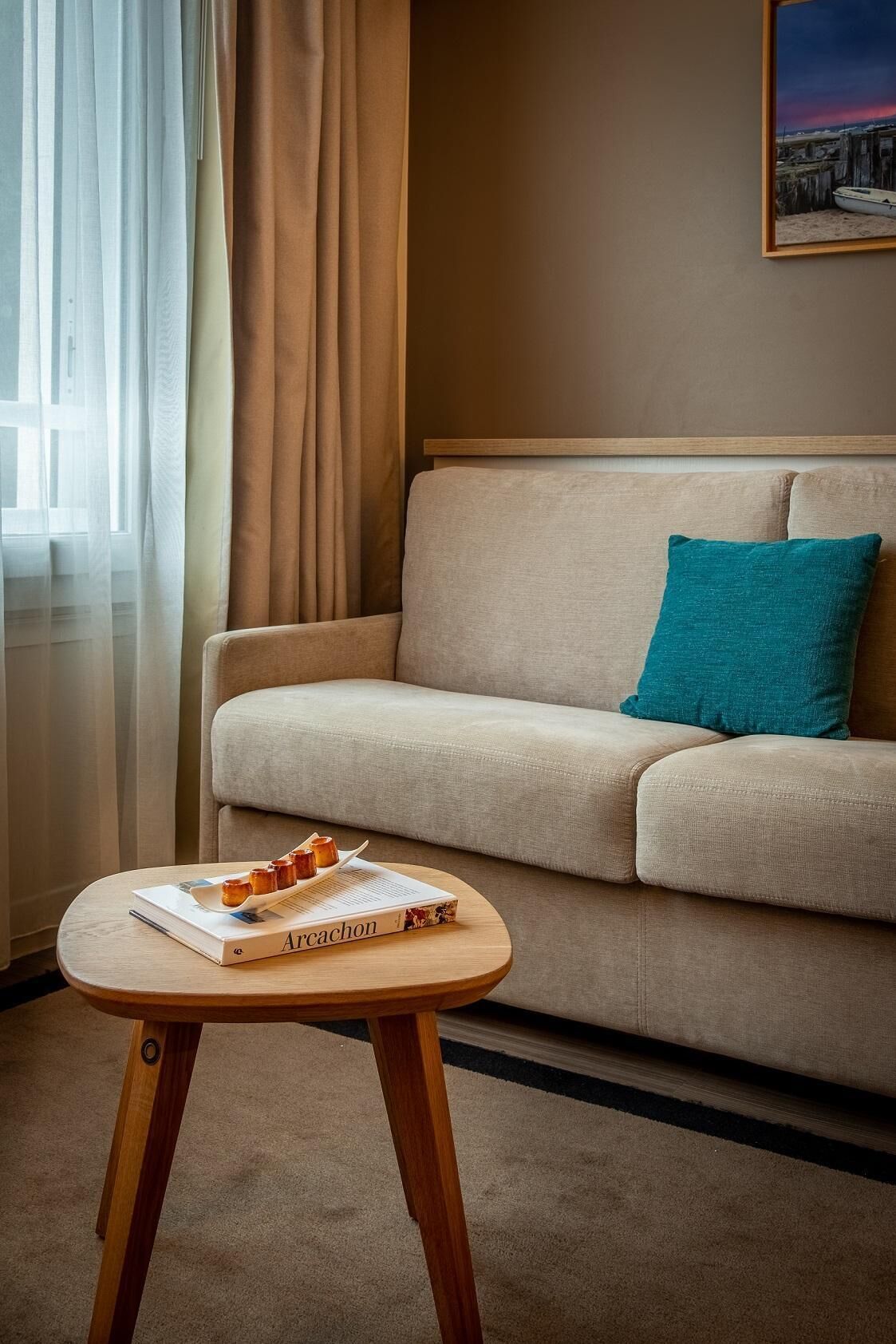 Un salon avec un canapé beige, un coussin turquoise, une petite table en bois sur laquelle repose un livre, et une fenêtre ornée de rideaux légers.