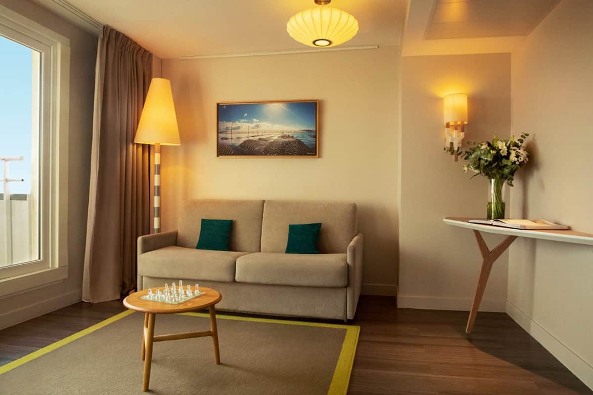 A cozy room with a sofa, a small table with a chessboard, a lamp, a painting, and a vase with flowers.