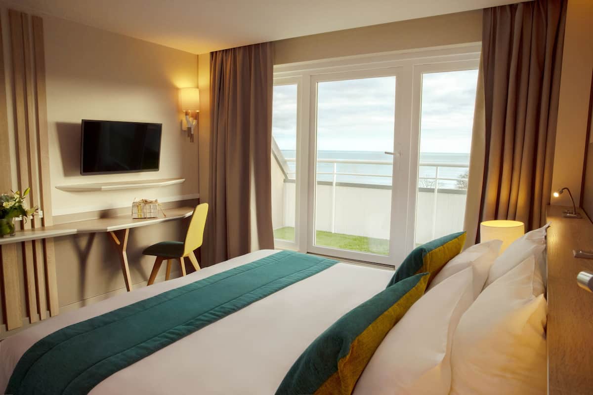 A hotel room with a bed, a desk with a chair, a television, and a view of the sea through large windows.