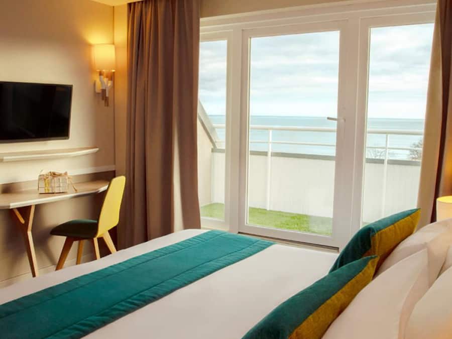 A hotel room with a bed, a desk, a chair, a TV, and a balcony with a view of the sea.