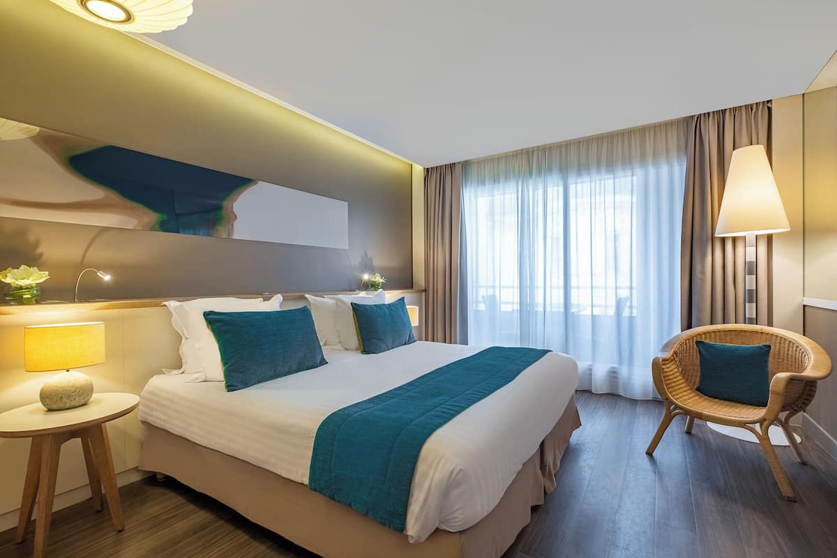 A hotel room with a bed, a chair, a lamp, and a window with curtains.