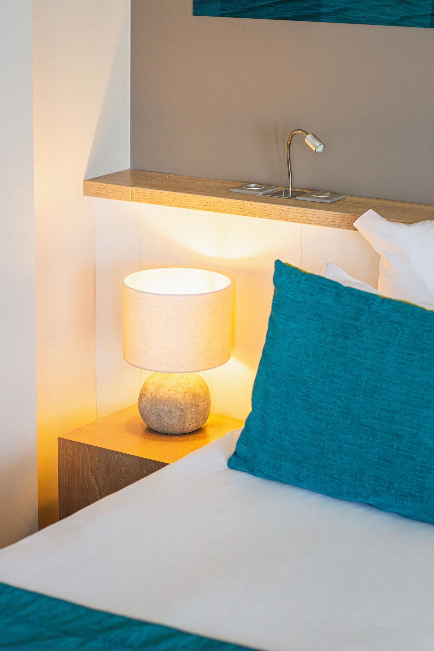 Habitación de hotel con cama, mesita de noche con lámpara, almohada azul y repisa de madera sobre la cama.