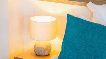Habitación de hotel con cama, mesita de noche con lámpara, almohada azul y repisa de madera sobre la cama.