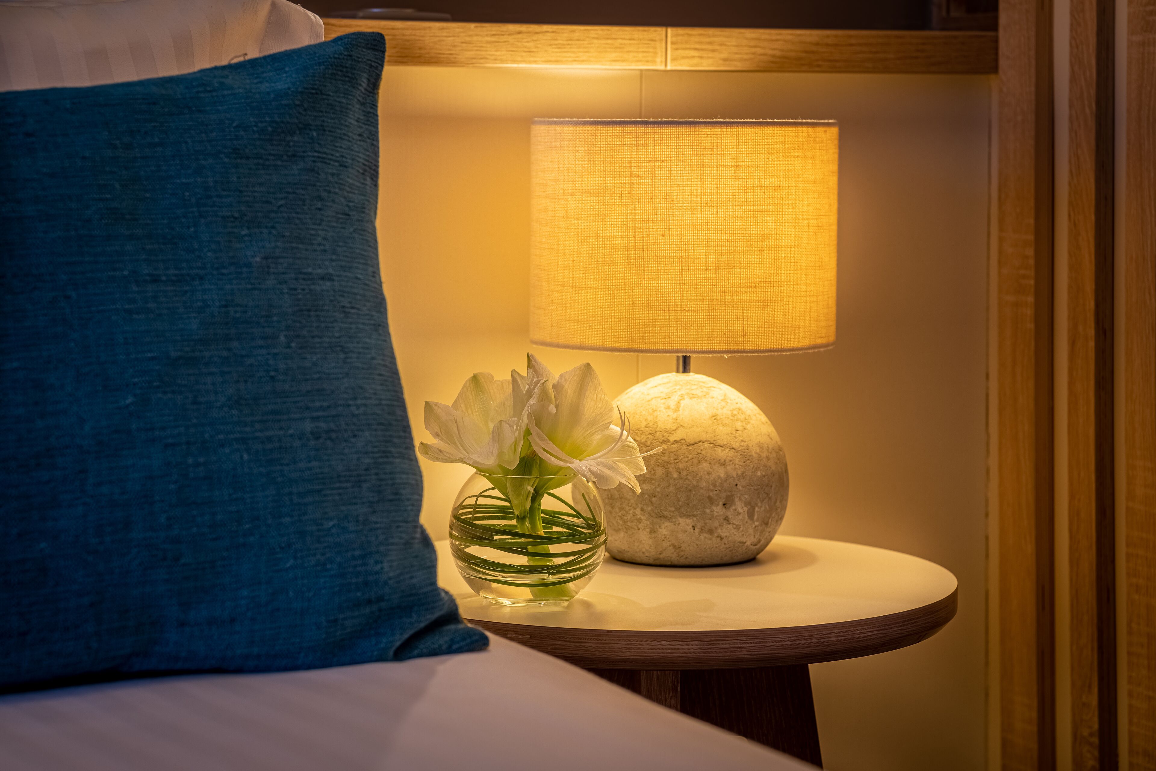 Una cama bien hecha con un cojín azul, una mesita de noche con una lámpara y un jarrón con flores.
