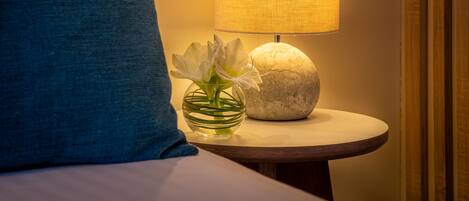 Una cama bien hecha con un cojín azul, una mesita de noche con una lámpara y un jarrón con flores.