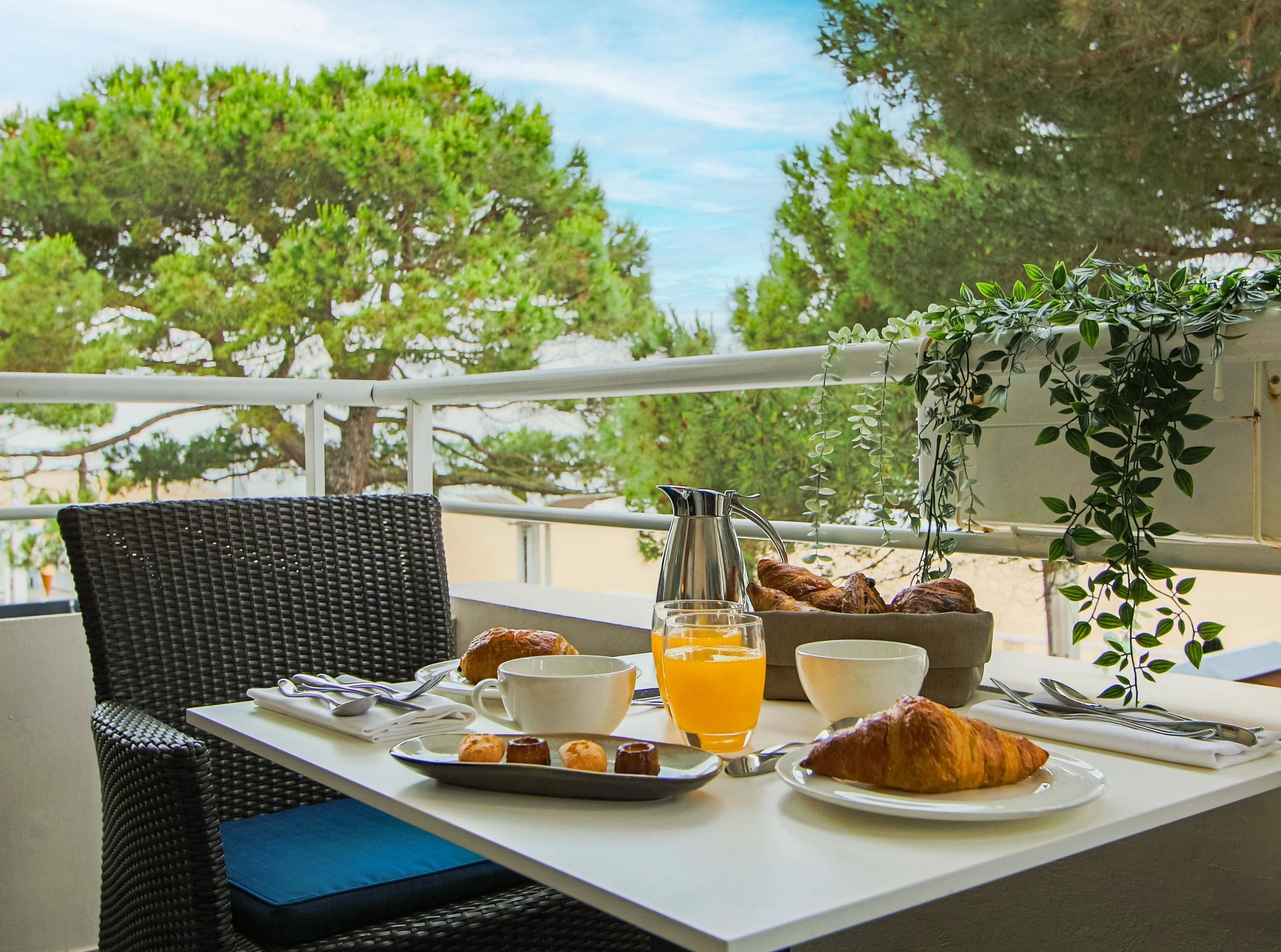 En morgenmadsopsætning på en balkon med en fletstol, et bord med croissanter, en kande, et glas appelsinjuice og en skål.