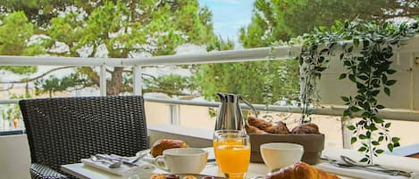 A breakfast setup on a balcony with a wicker chair, a table with croissants, a pitcher, a glass of orange juice, and a bowl.