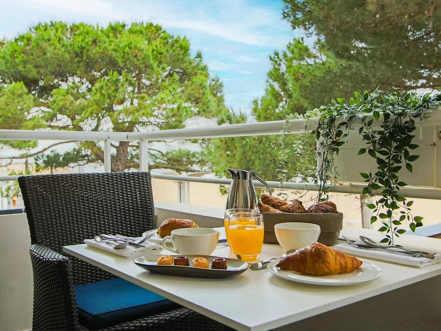 A breakfast setup on a balcony with a wicker chair, a table with croissants, a pitcher, a glass of orange juice, and a bowl.