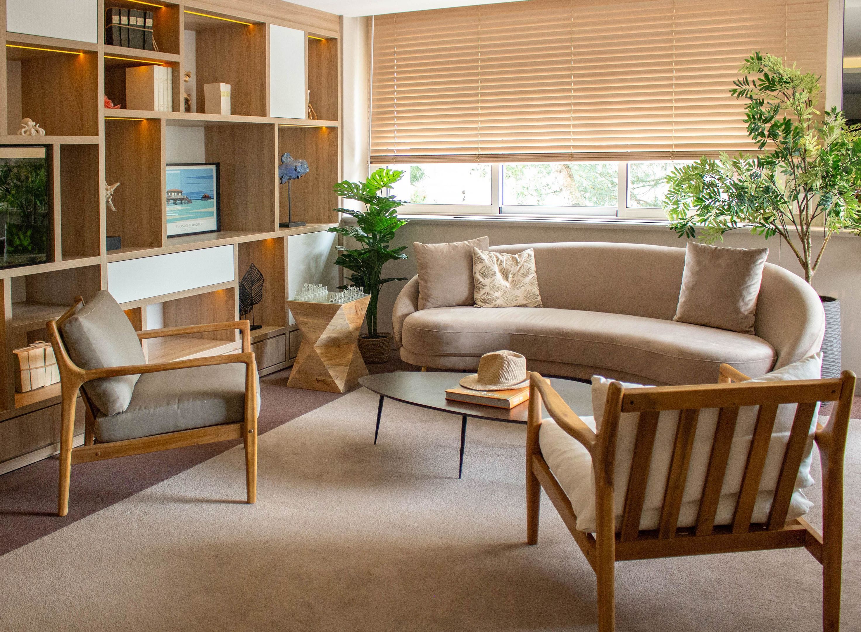 Een moderne woonkamer met een beige bank, houten stoelen, een salontafel en een boekenplank met diverse objecten.