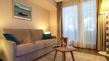 A hotel room with a sofa, a table with a bottle and glasses, a painting on the wall, and a window with curtains.