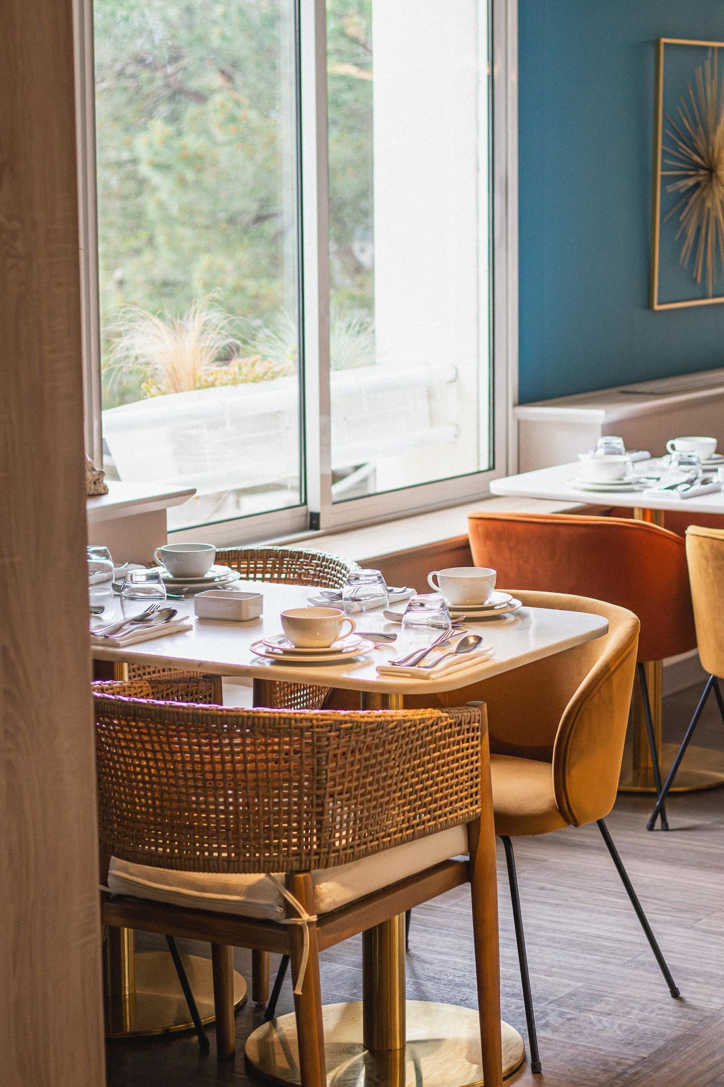 A dining area with a table set for two, wicker chairs, and a view of greenery through large windows.