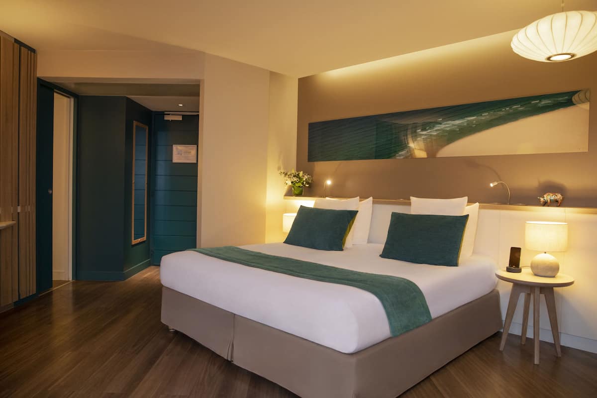 A modern hotel room with a large bed, a bedside table with a lamp, and a wall-mounted artwork.
