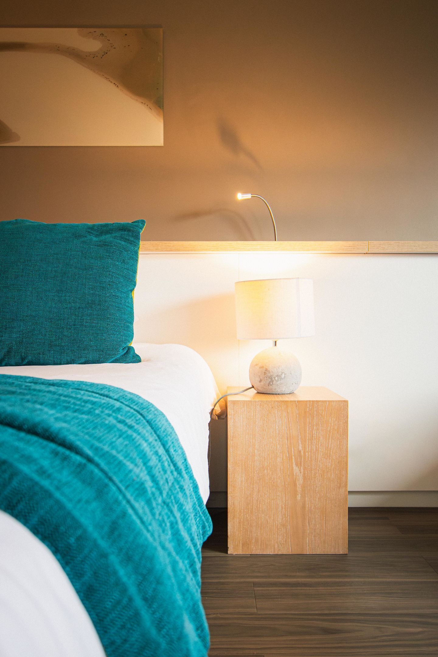 A neatly made bed with a teal pillow and a white blanket, a bedside table with a lamp, and a wooden nightstand.