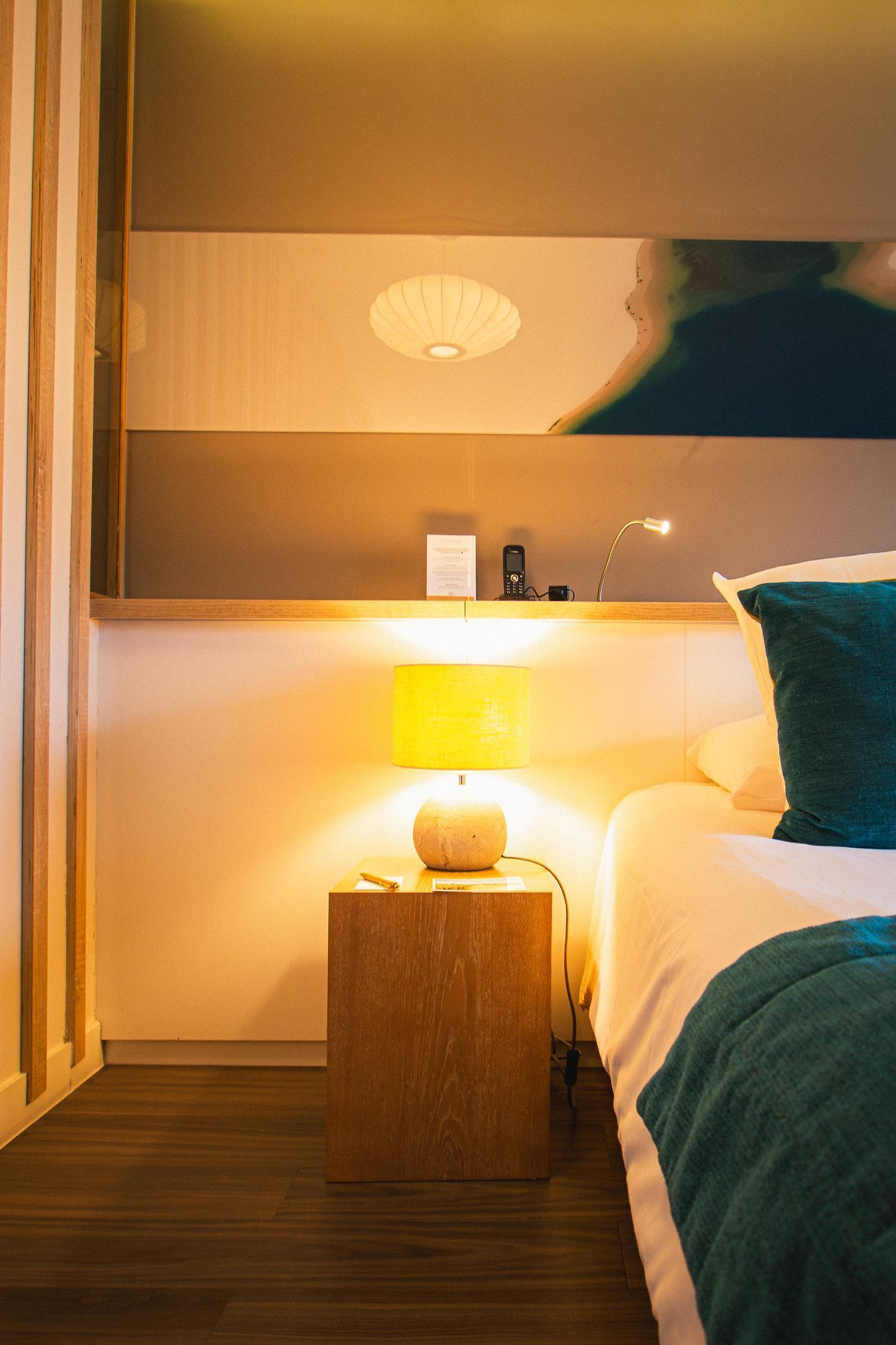 A hotel room with a bed, a bedside table with a lamp, a wall-mounted phone, and a small wooden nightstand.
