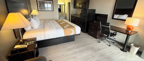 Habitación de hotel con cama, mesitas de noche, escritorio, silla, espejo, televisor y kitchenette.