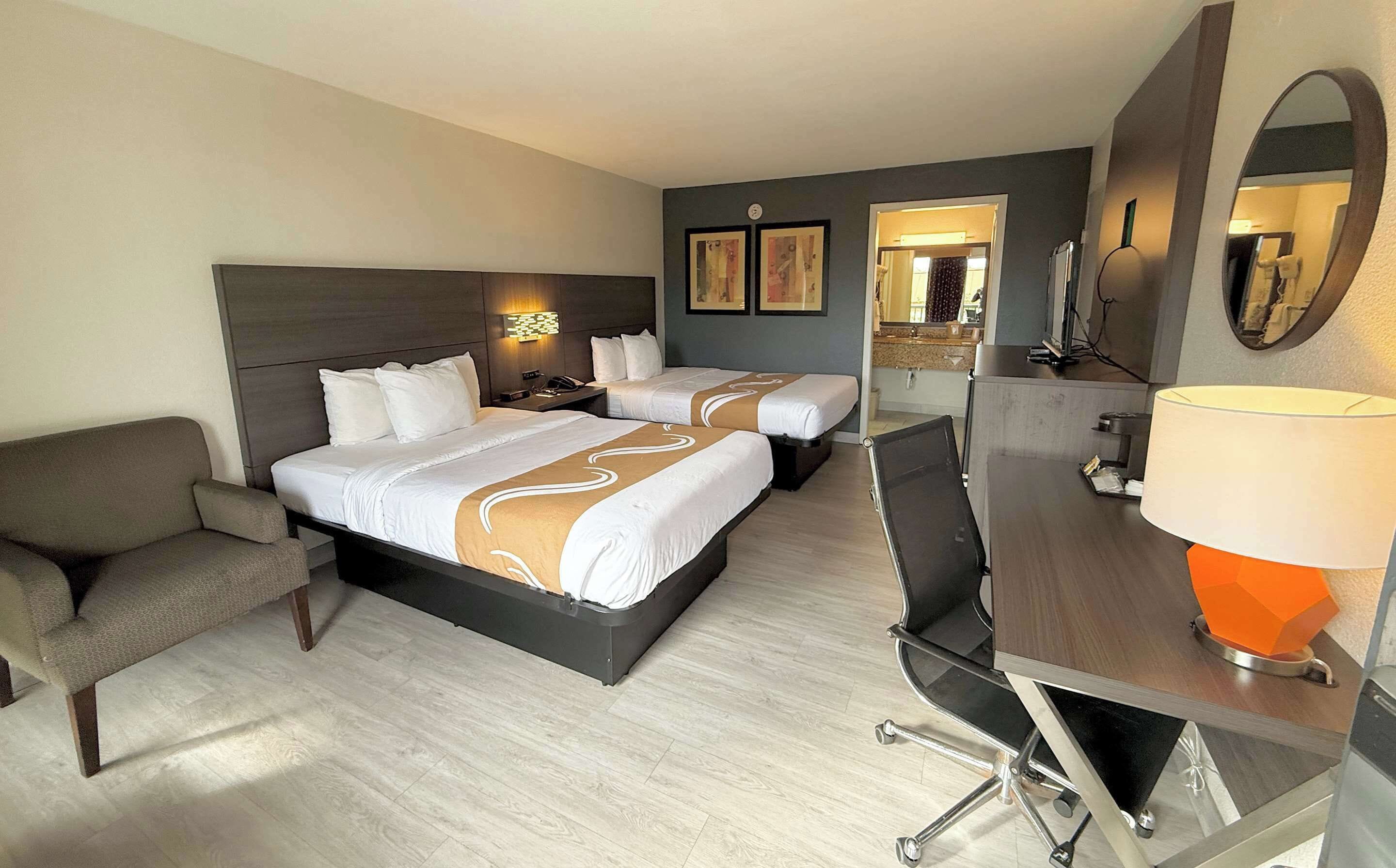 Ett hotellrum med två sängar, ett skrivbord, en stol, en lampa och en spegel.