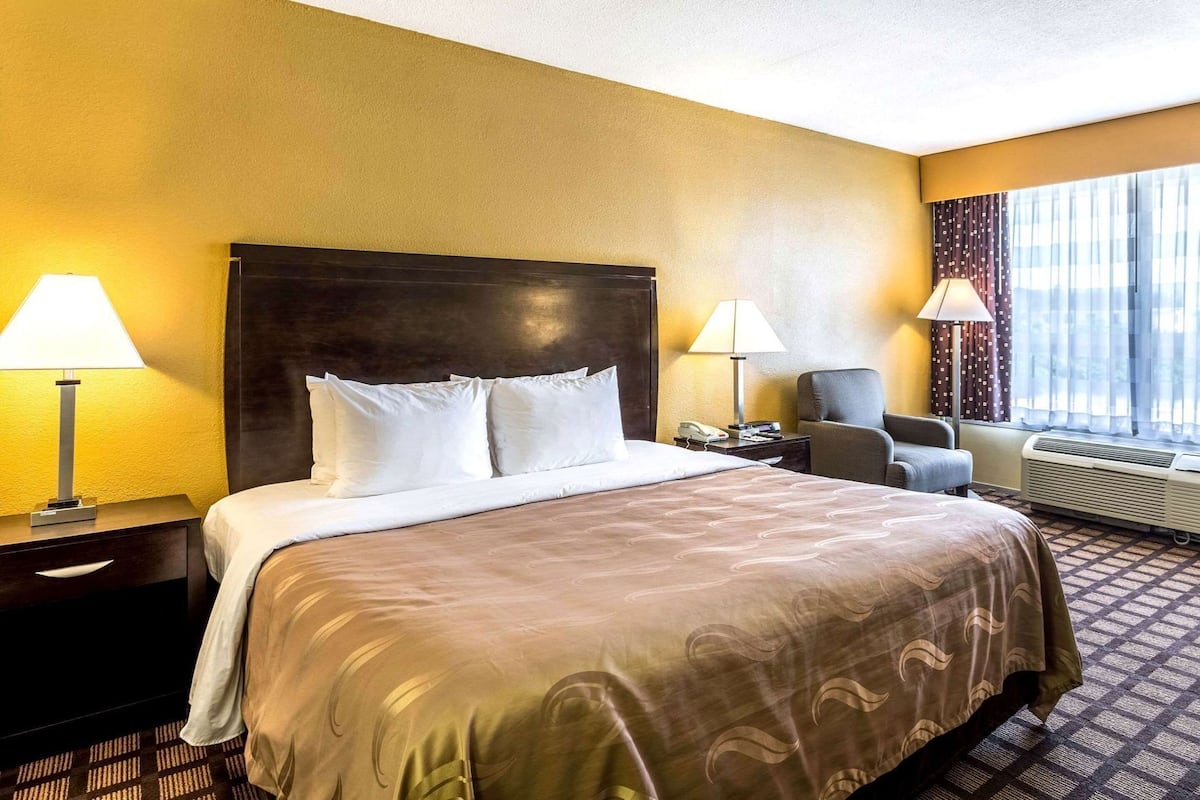 A hotel room with a large bed, two bedside lamps, a chair, a desk with a phone, and a window with curtains.