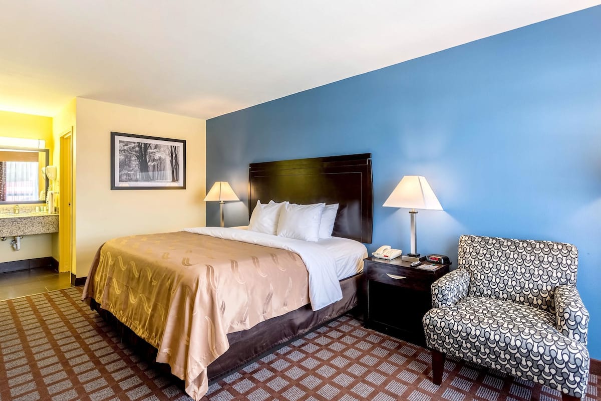 A hotel room with a bed, bedside lamps, a chair, a nightstand, and a framed picture on the wall.