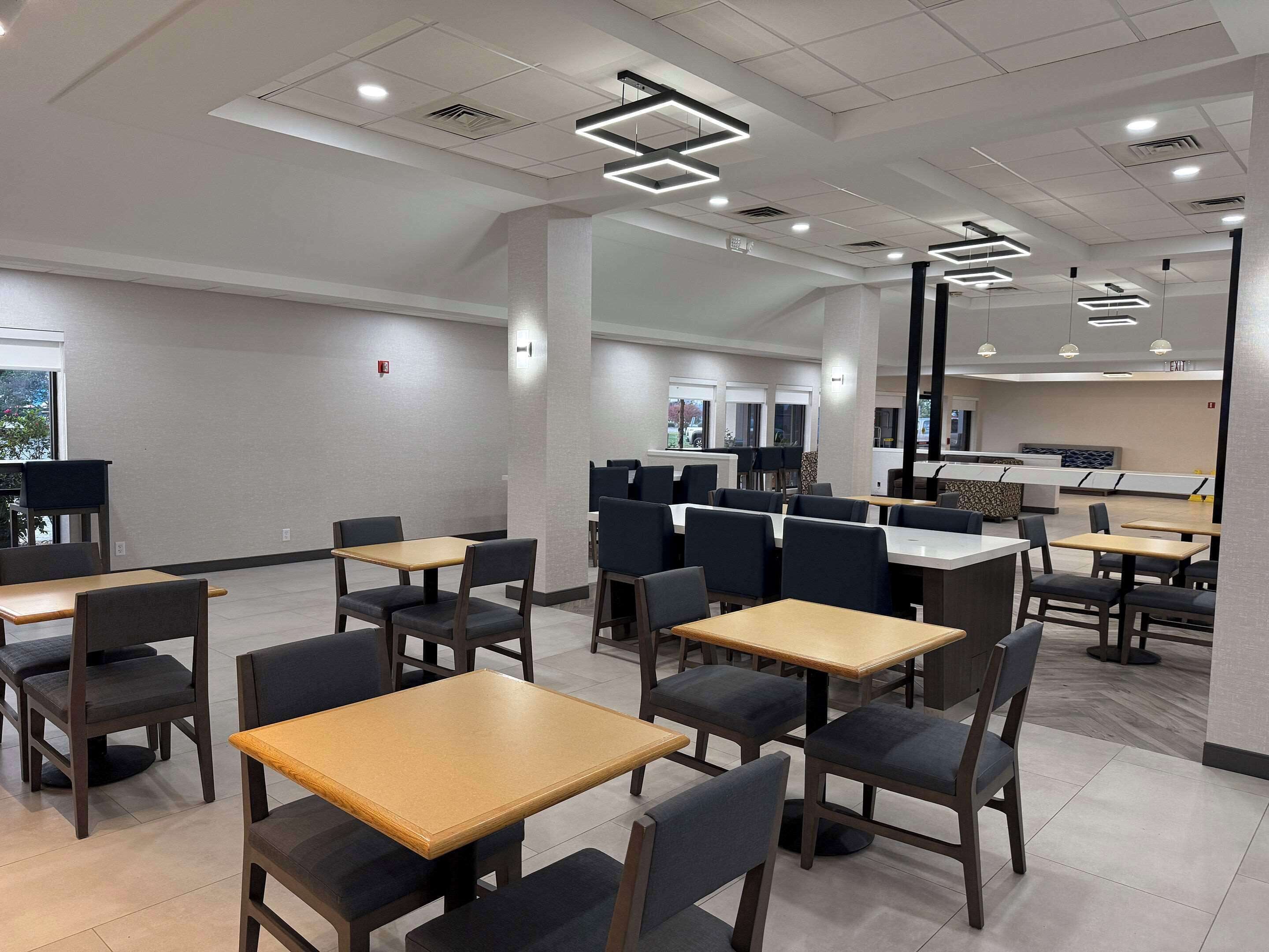 A modern dining area with rectangular tables and chairs, recessed ceiling lighting, and a view of the outside through large windows.