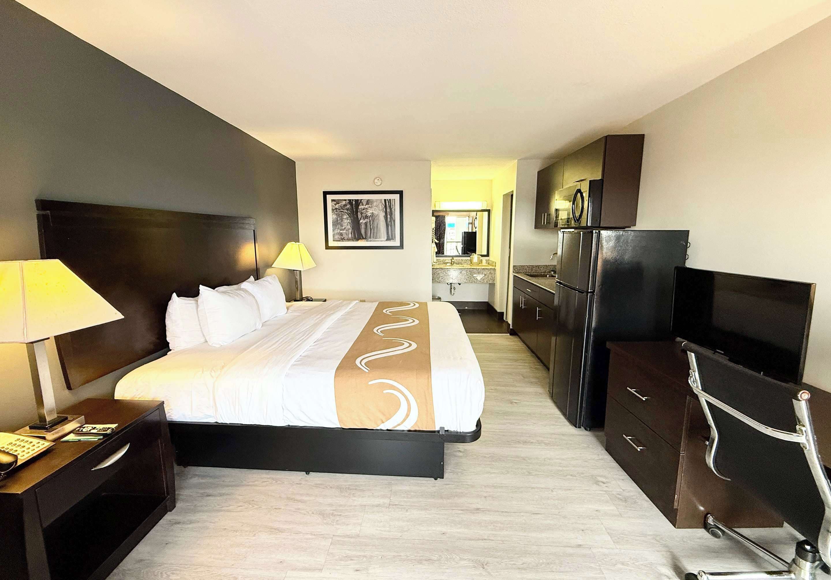 A hotel room with a bed, a desk with a lamp, a TV, a dresser, and a kitchen area with a refrigerator and microwave.