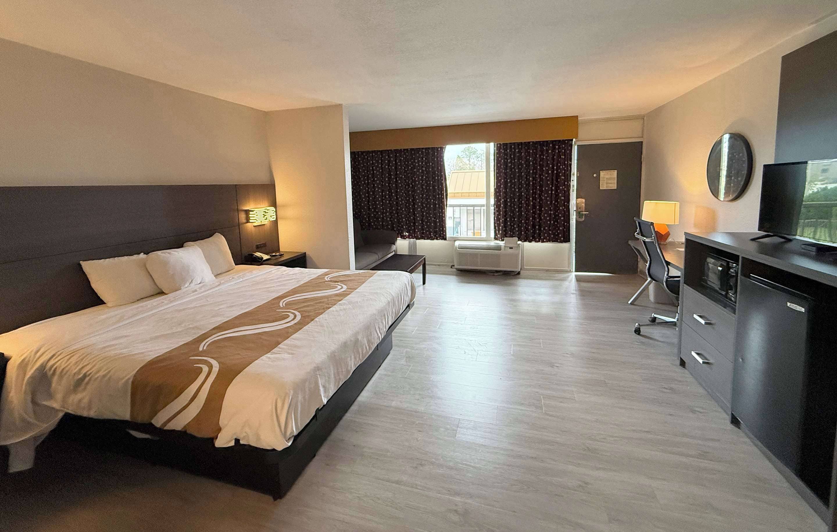 A hotel room with a large bed, a desk with a computer, a TV, and a window with curtains.