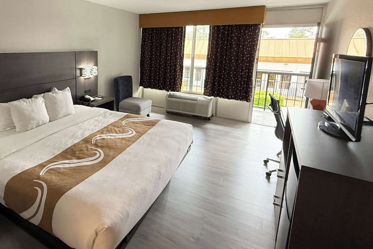A modern hotel room with a large bed, a desk with a computer, a TV, and a window with curtains.