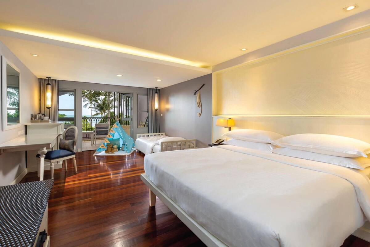 A hotel room with a large bed, a desk, a chair, a small table with a decorative item, and a view of palm trees through the window.