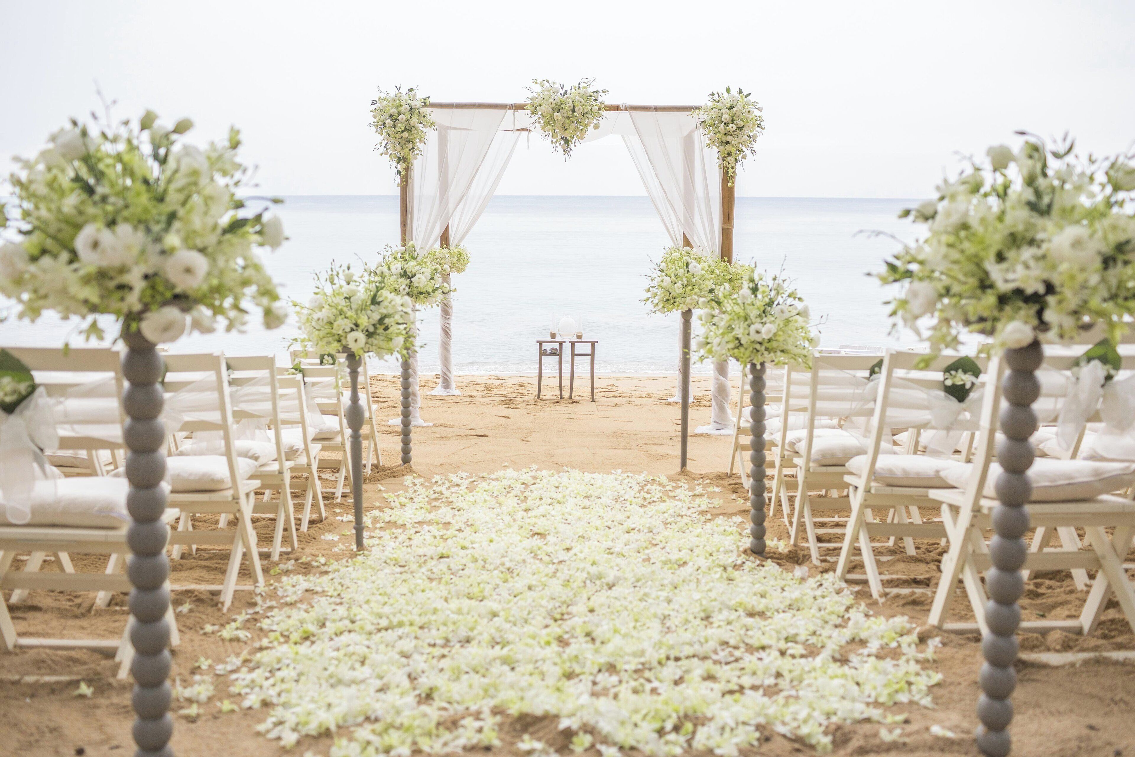 A beach wedding setup with a white aisle, chairs, and floral decorations.