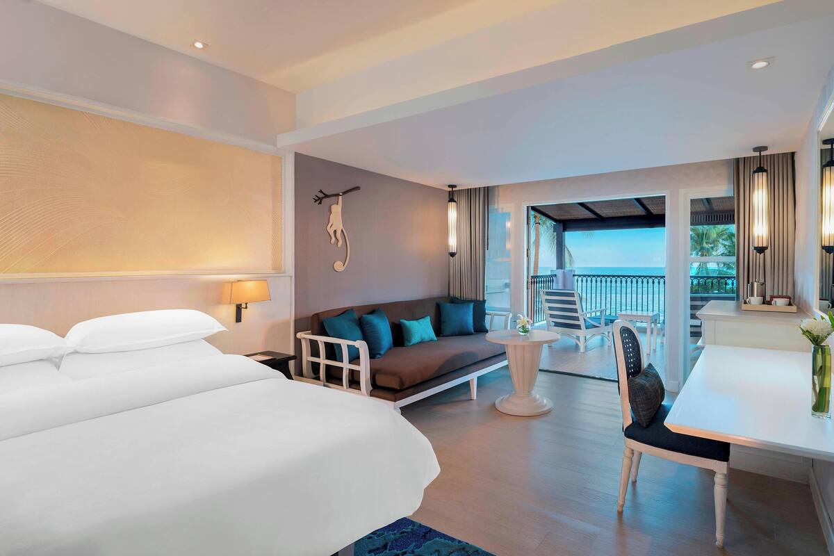 A hotel room with a large bed, a sofa with teal cushions, a dining table, and a view of the outdoors.