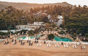 A resort with a swimming pool surrounded by lounge chairs and umbrellas, set against a backdrop of palm trees and buildings.