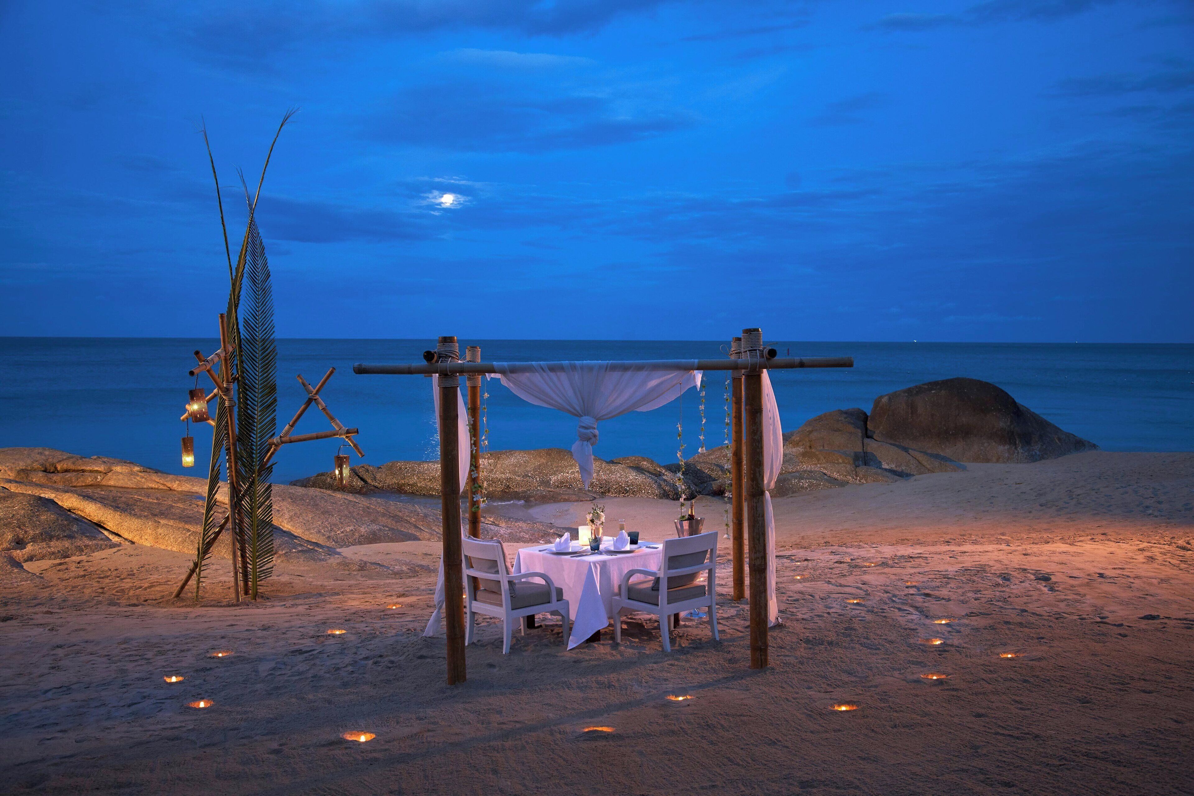 Un dîner organisé sur la plage, avec une table et des chaises placées sous un auvent, entourées d’éléments décoratifs et offrant une vue sur la mer.