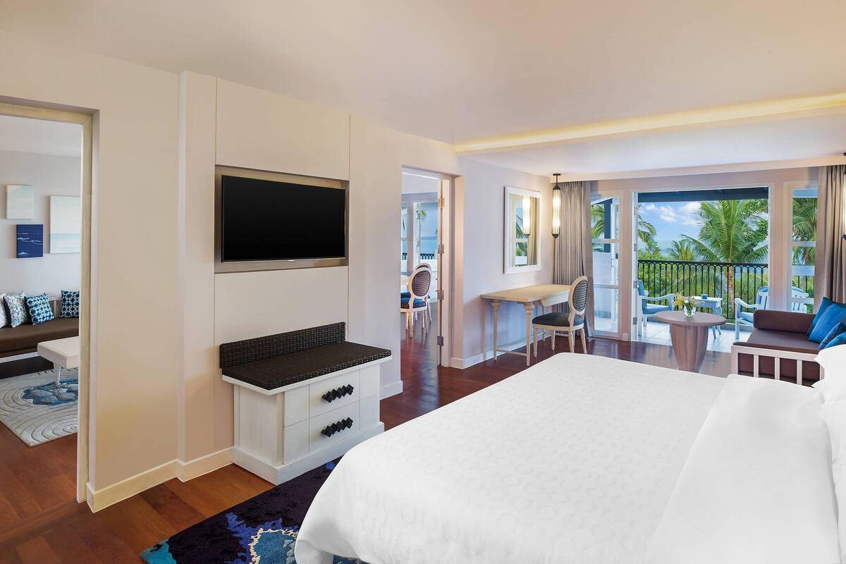 A hotel room with a large bed, a TV, a desk with a chair, and a balcony with outdoor seating.