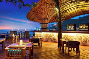 Une terrasse en bois avec des tables et des chaises, un coin bar avec des bouteilles, et une vue sur l’océan au crépuscule.