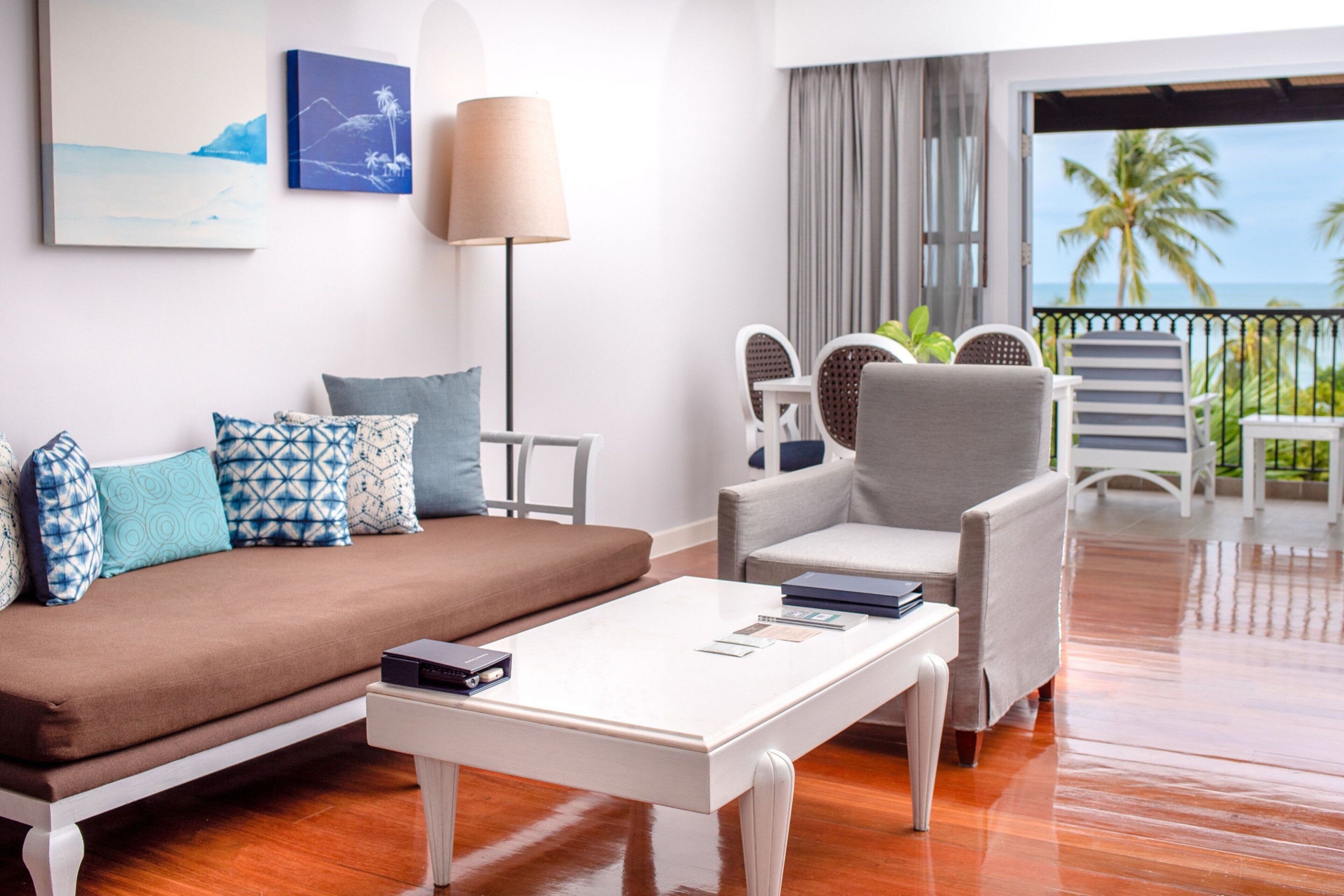 A modern hotel room with a sofa, armchairs, a coffee table, and a balcony with a view of palm trees and the sea.
