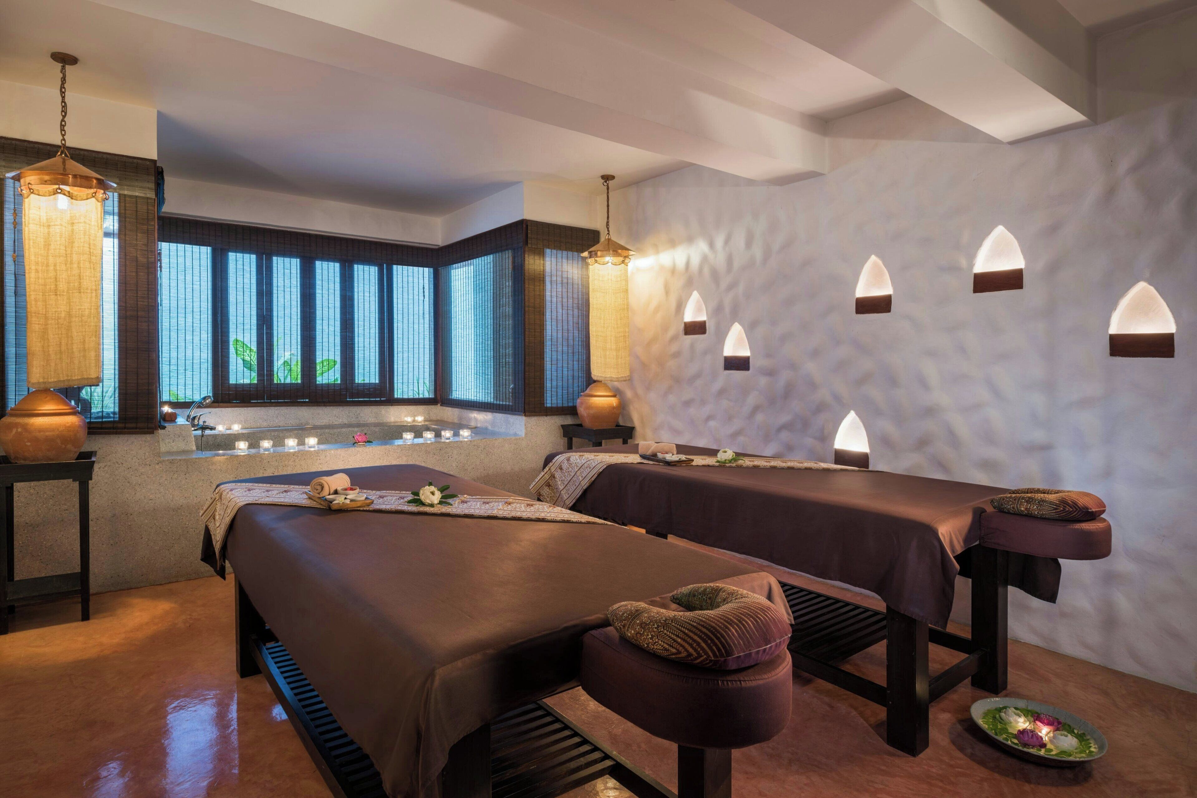 A spa room with two massage tables, a large window with vertical blinds, and a wall-mounted light fixture.