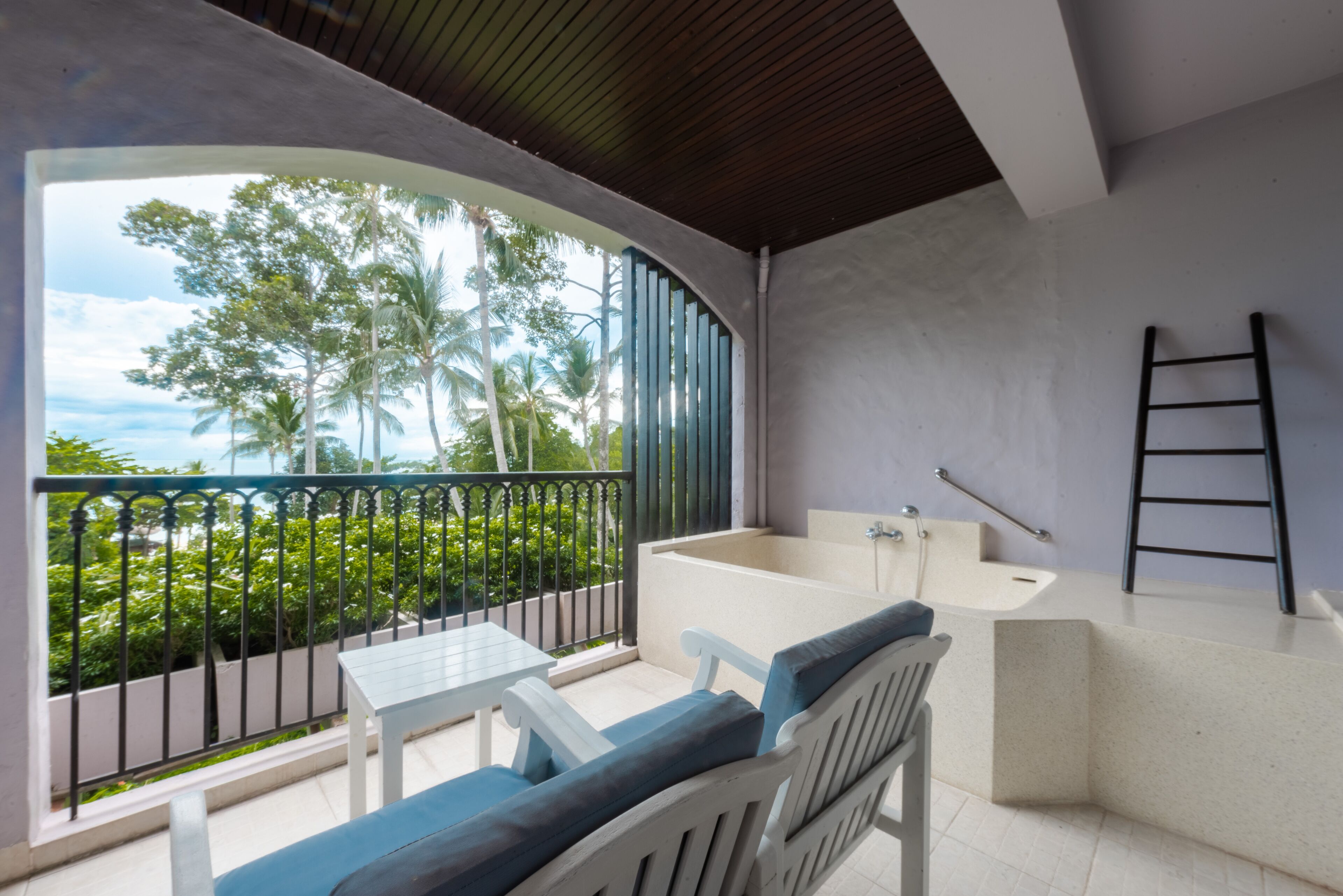 A balcony with a view of palm trees and the sea, a bathtub, and a white chair with blue cushions.