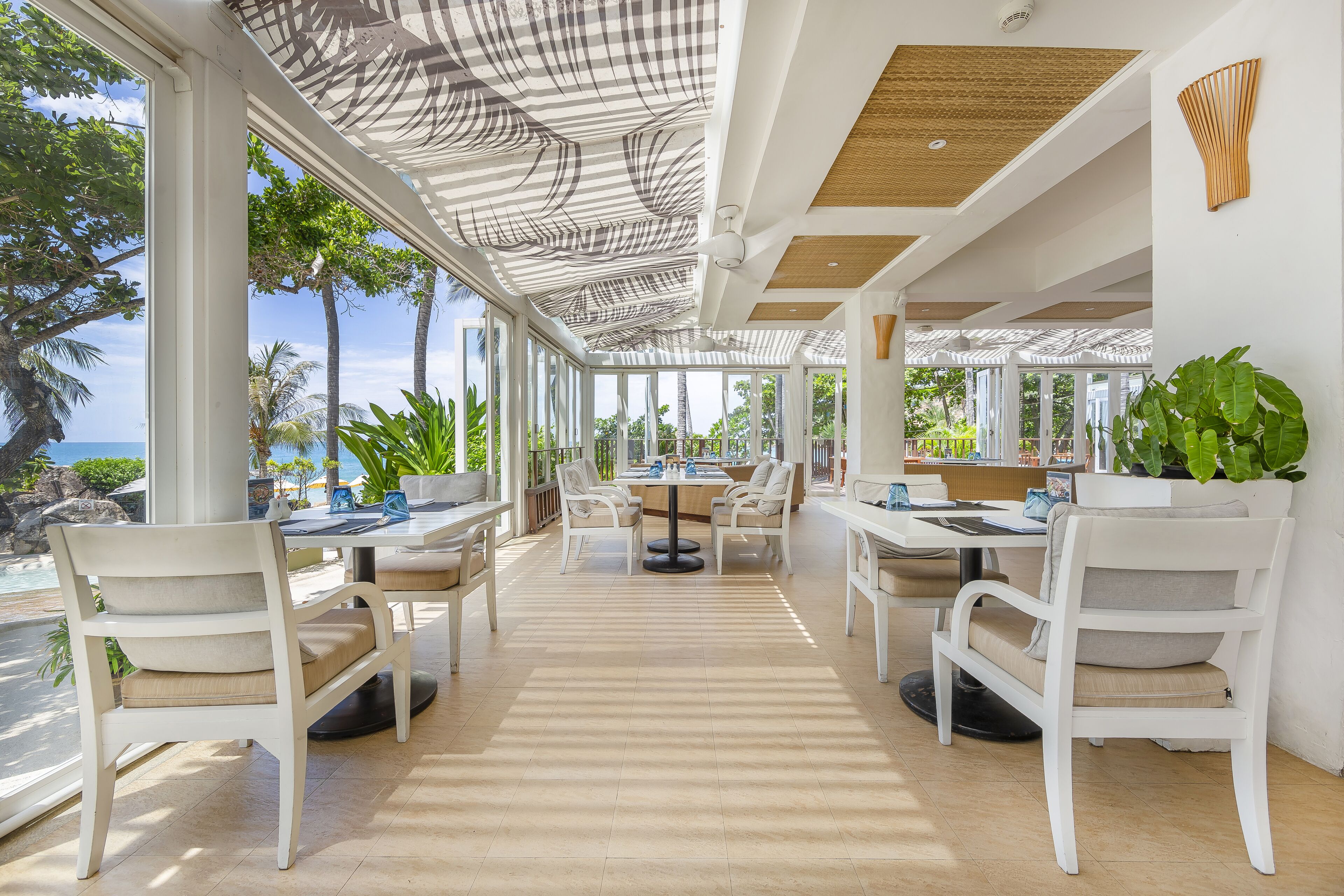 A spacious outdoor dining area with white furniture, large windows, and a view of the ocean.