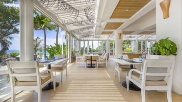 A spacious outdoor dining area with white furniture, large windows, and a view of the ocean.