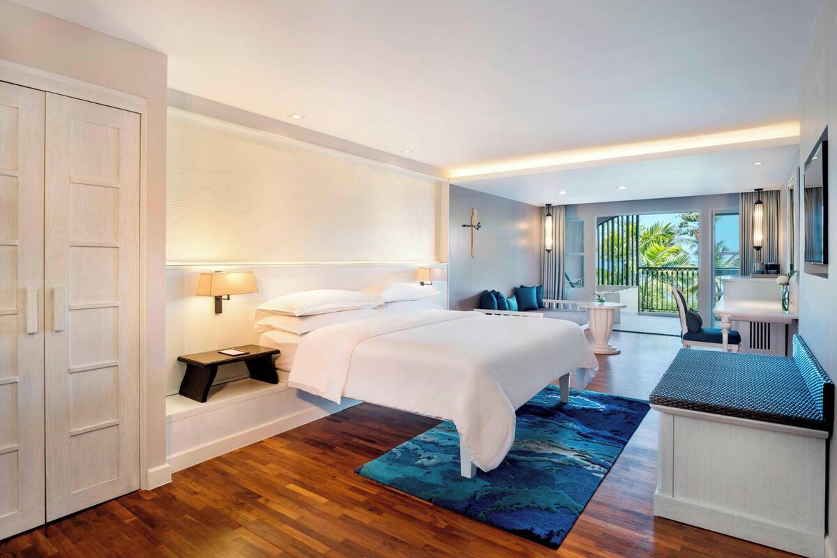 A modern hotel room with a large bed, a small desk, a balcony with a view, and a large window.