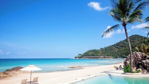 Une plage avec une eau turquoise et limpide, un rivage sablonneux, une piscine, des chaises longues et un palmier.