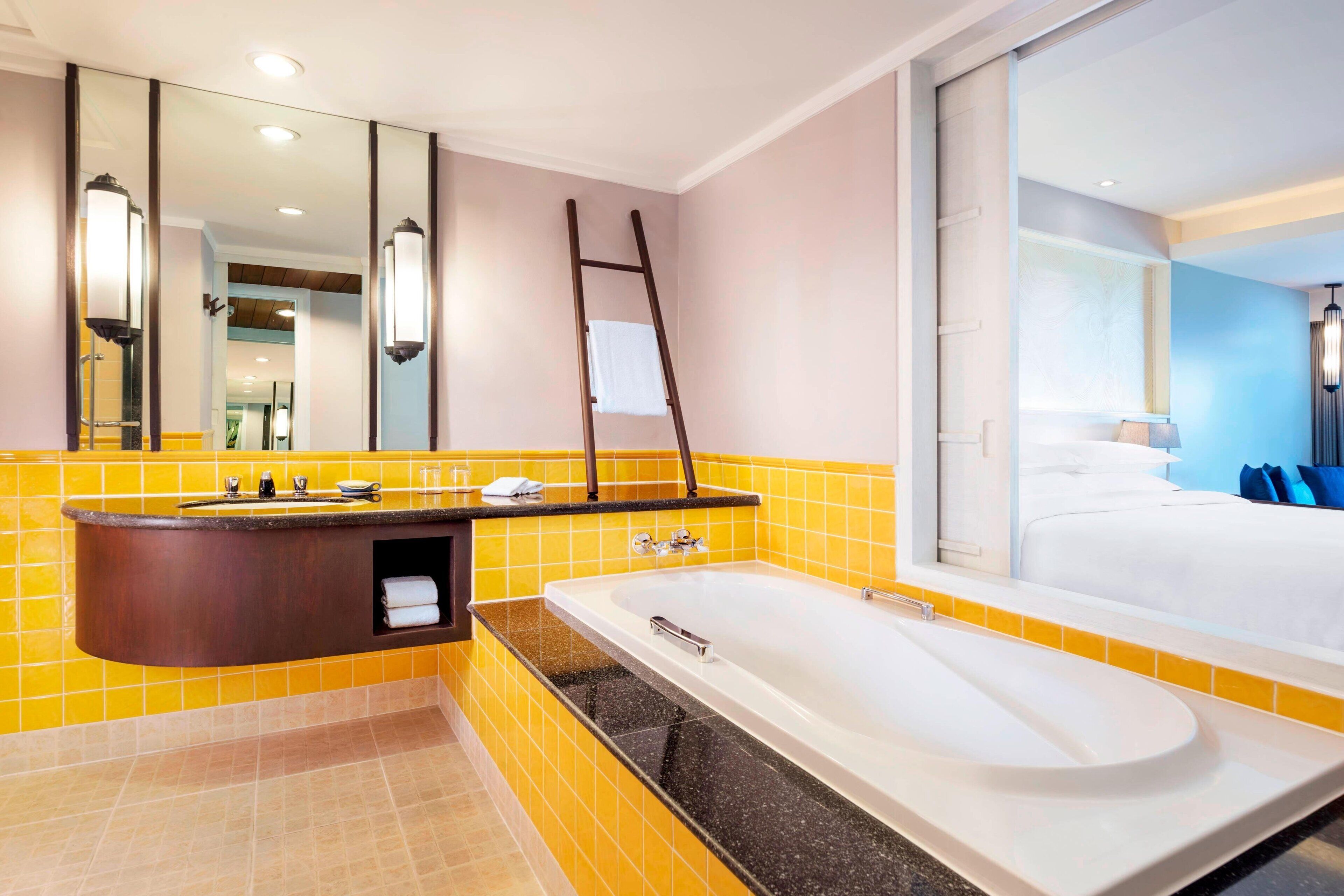 Een moderne badkamer met een groot bad, een wastafel met spiegel, en een raam met uitzicht op een slaapkamer.