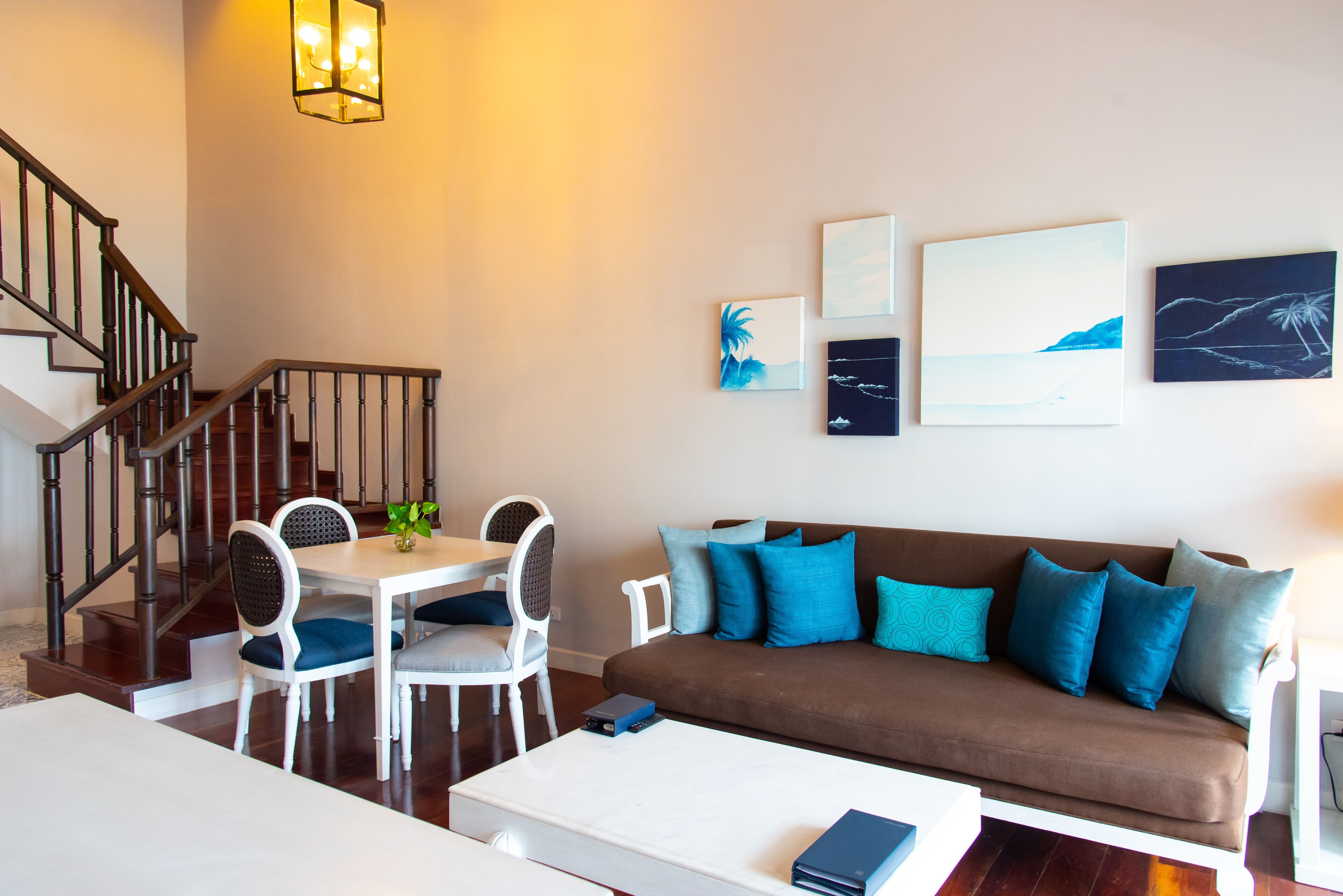 Una sala de estar con un sofá marrón, una mesa de centro blanca y un comedor con una mesa y sillas blancas. Hay cuadros en la pared y una escalera con barandilla.