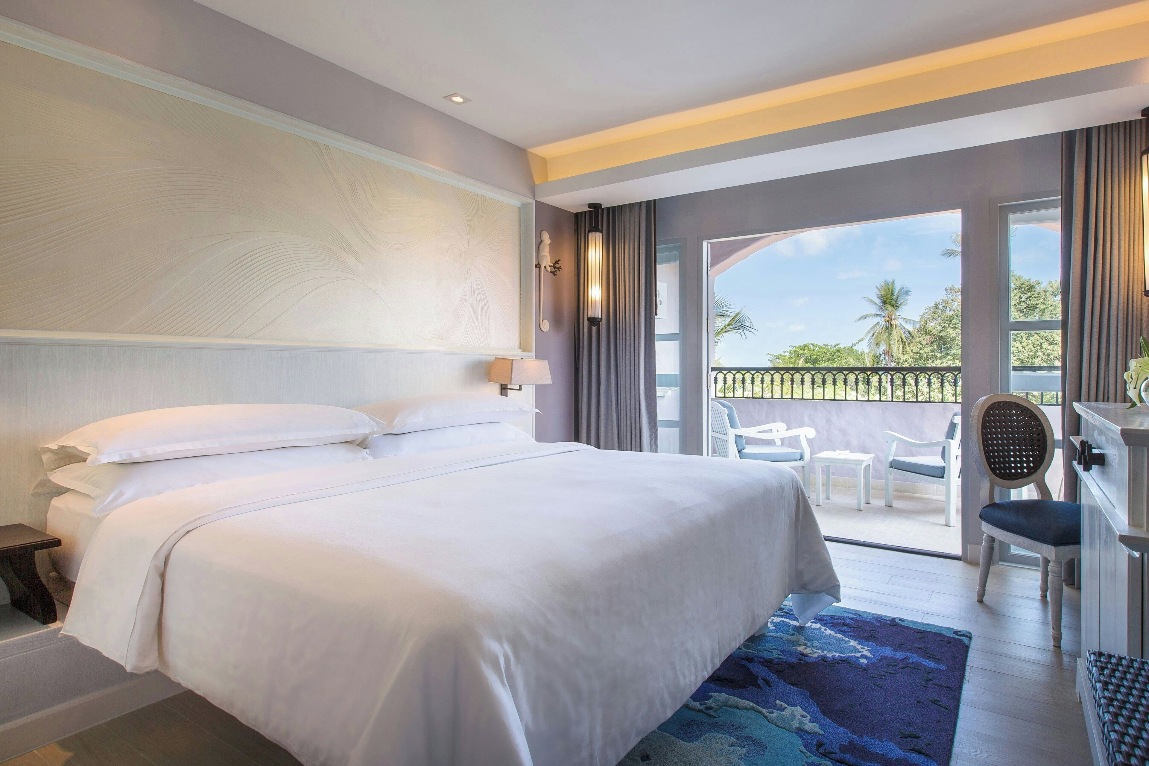 A hotel room with a large bed, a desk with a chair, a balcony with a view of palm trees, and a window with curtains.