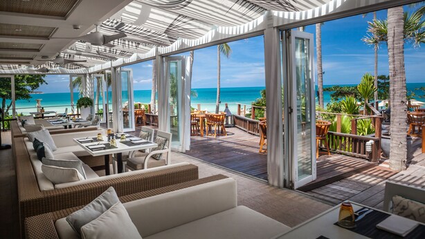 A modern outdoor lounge area with wicker furniture, glass doors leading to a deck, and a view of the ocean. - Sheraton Samui Resort (Koh Samui)