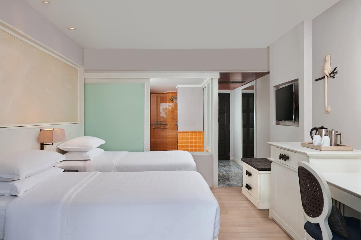 A hotel room with two beds, a desk, a TV, and a bathroom visible through an open door.