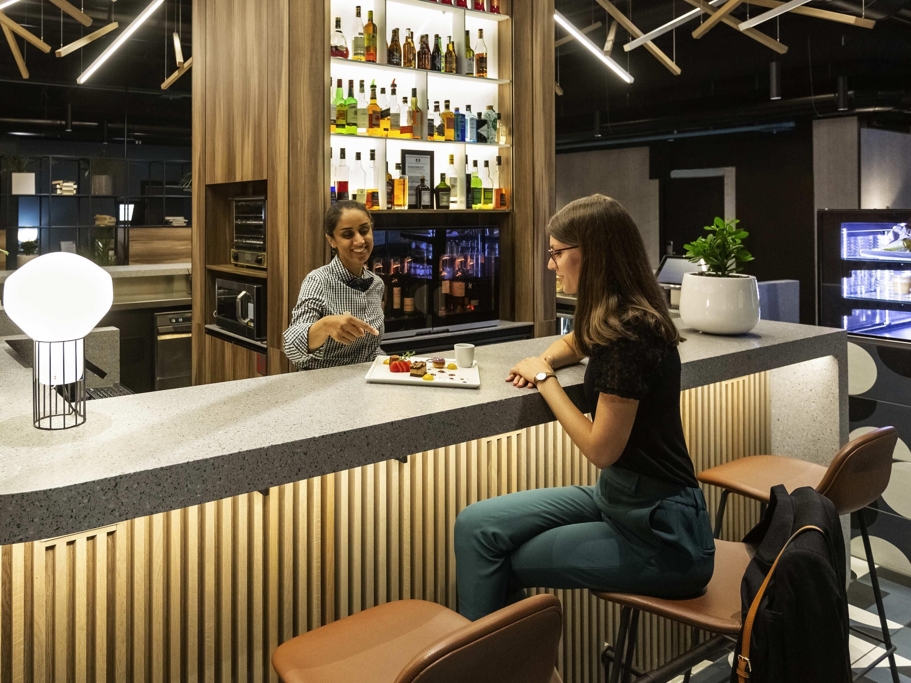 Un espace bar comprenant un comptoir, des tabourets, une plante en pot et une étagère avec différentes bouteilles.